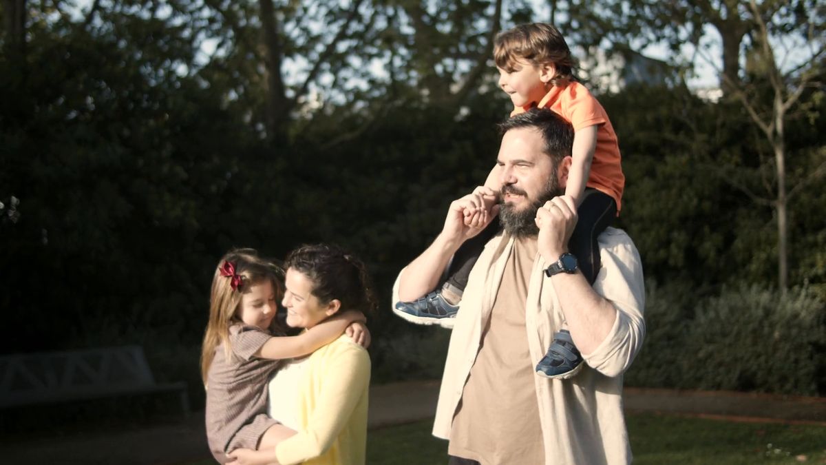A dad with his young son on his shoulders, and a mum hugging their daughter, on a sunny afternoon in the park
