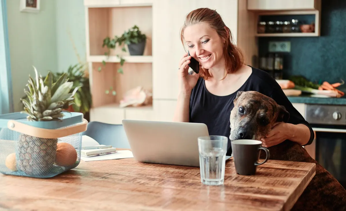 A smiling woman on the phone at her laptop in the kitchen, her dog beside her