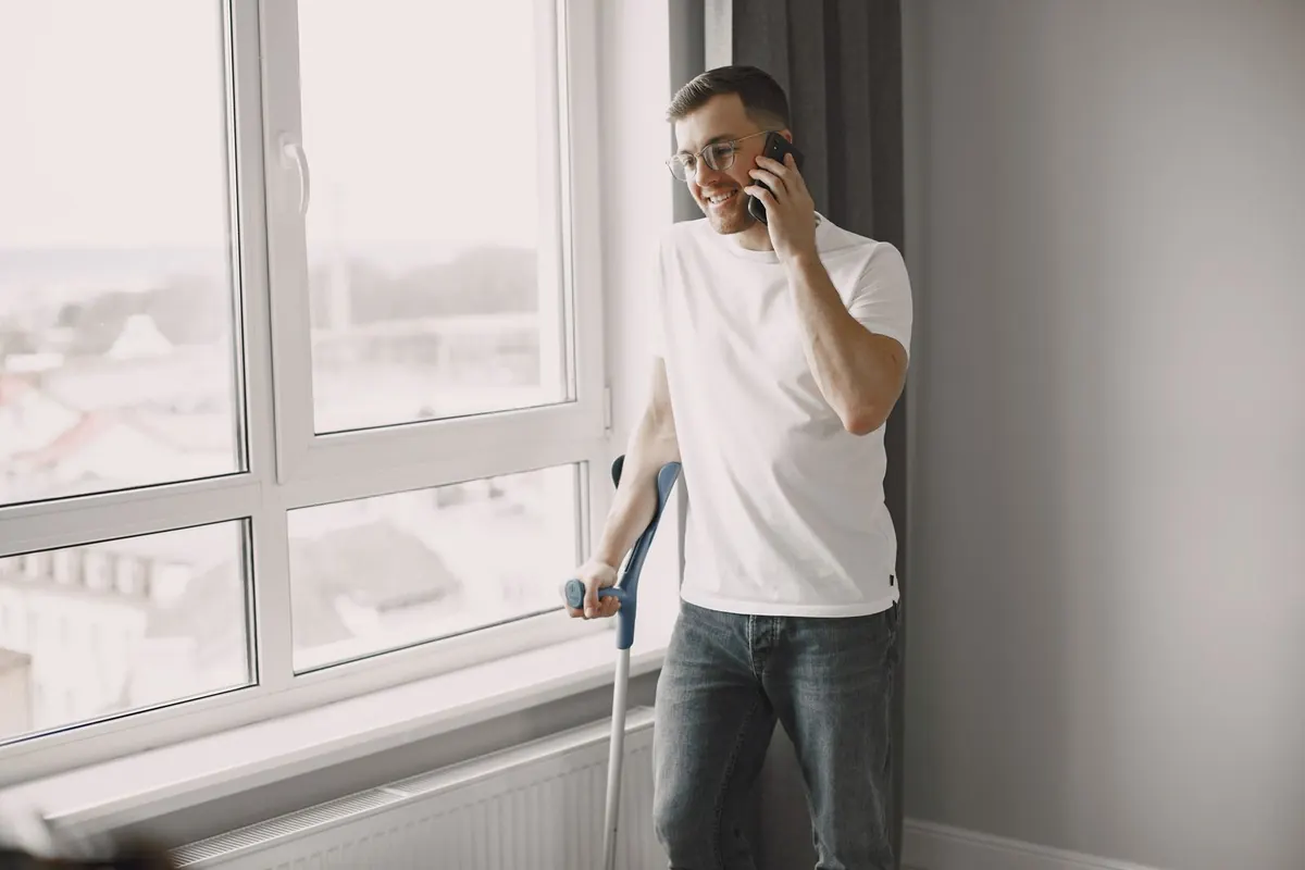 A young man on a crutch at home, on the phone by the window